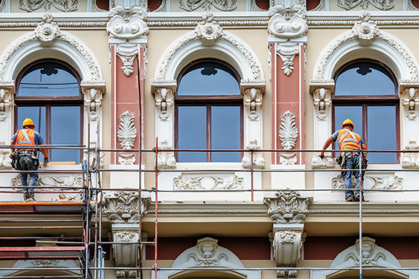 Das Bild zeigt zwei Bauarbeiter auf einem Gerüst bei der Sanierung einer reich verzierten historischen Hausfassade.